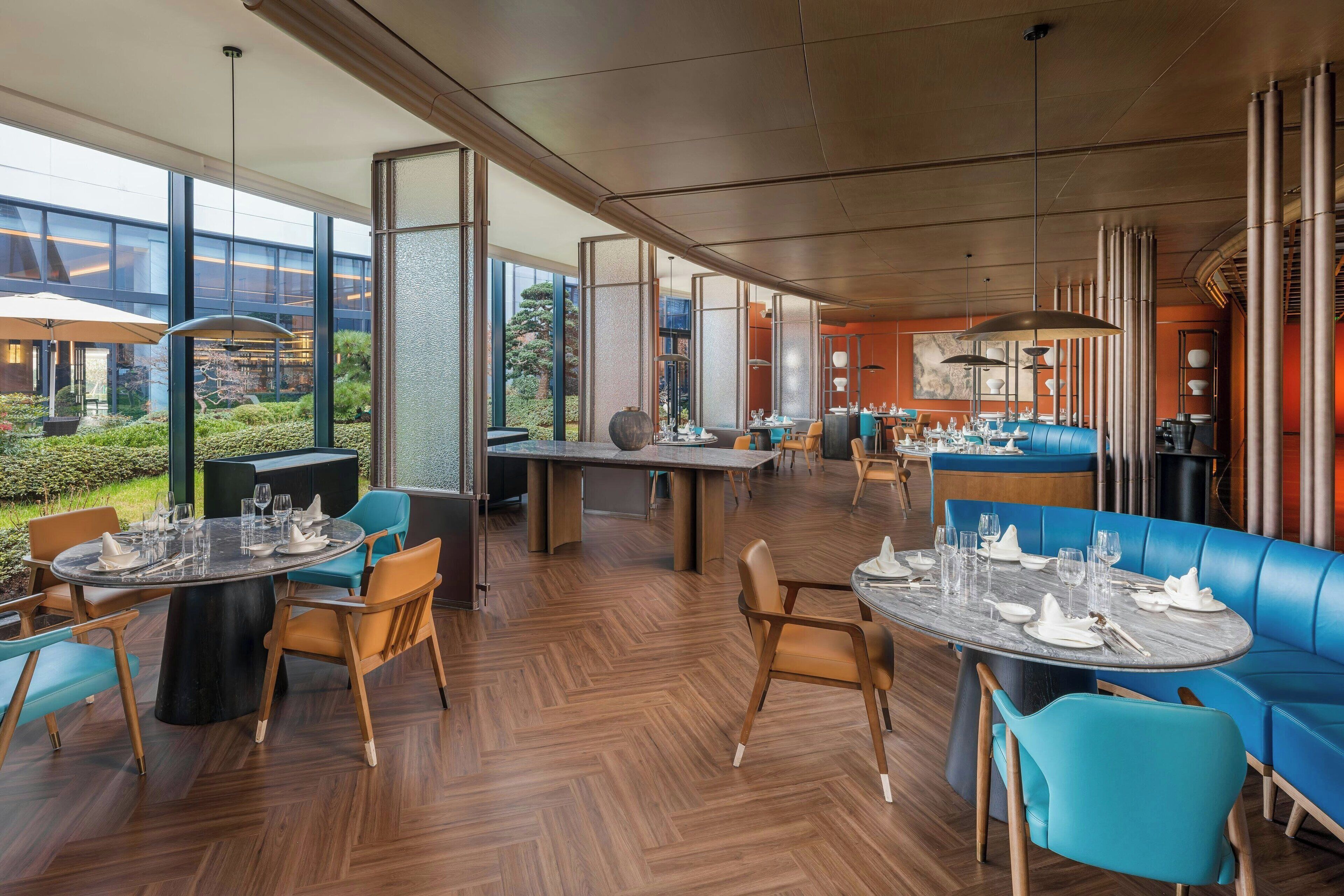 A modern restaurant with round tables and blue seating, large windows, and a view of greenery.