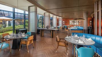 A modern restaurant with round tables and blue seating, large windows, and a view of greenery.