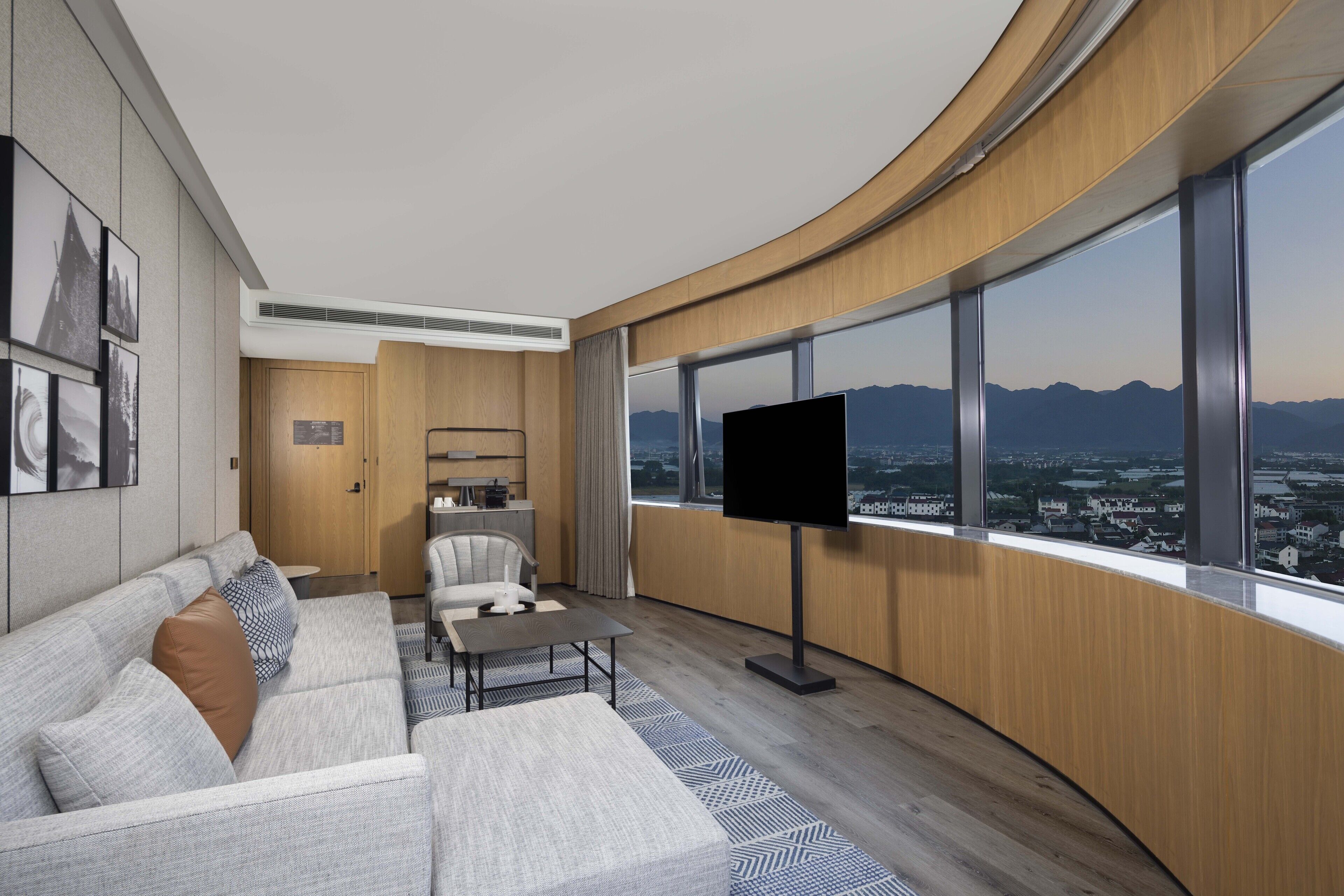 A modern hotel room with a curved wooden wall, a large TV, a sofa, a chair, and a view of a cityscape.