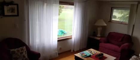 A living room with a wooden coffee table, a red armchair, a floral sofa, and a window with sheer curtains.