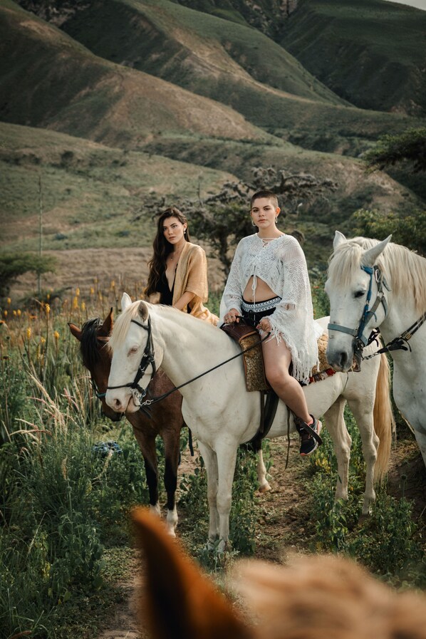 Two individuals riding horses on a dirt path surrounded by greenery and hills.