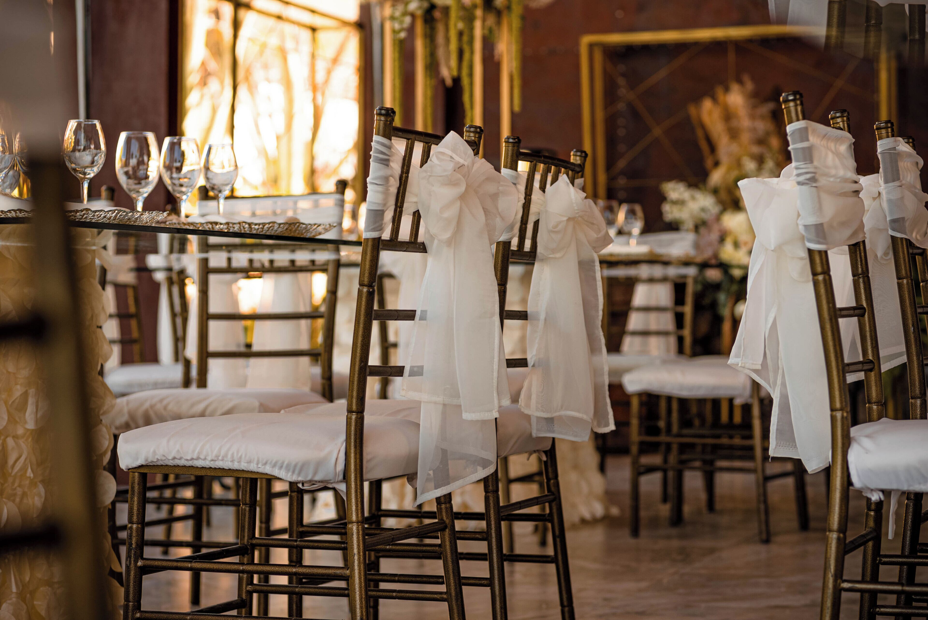 A row of elegantly arranged chairs with white cushions and sheer fabric bows, set against a warm interior with a decorative mirror and framed artwork.