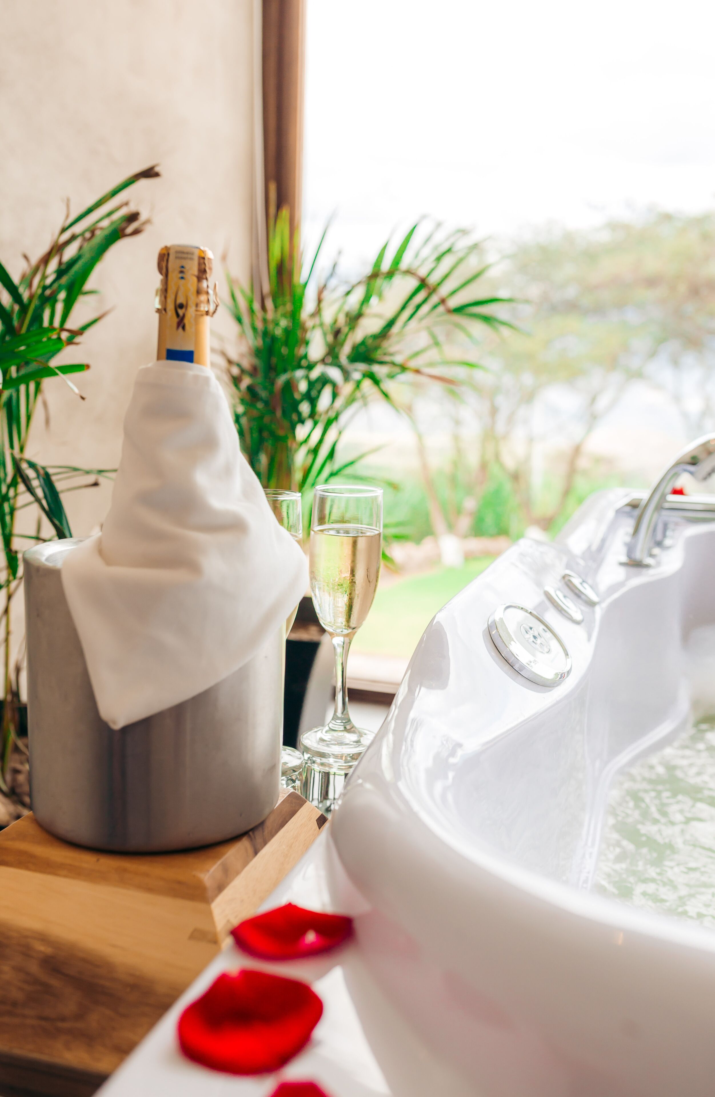 A bathtub with rose petals, a bottle of champagne, and glasses on a wooden table.