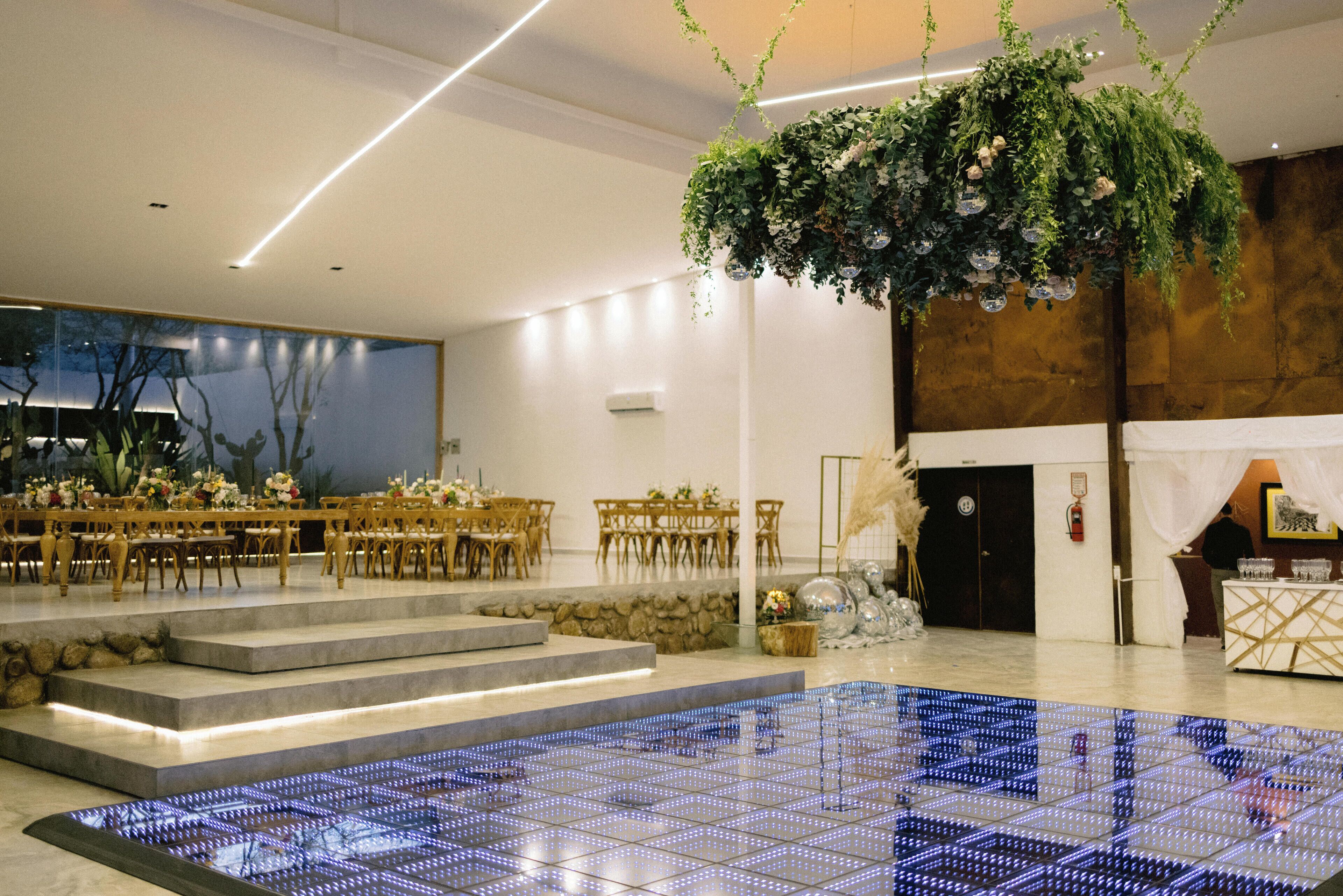 A modern hotel lobby with a large, illuminated dance floor, a reception desk, and a staircase leading to a dining area.