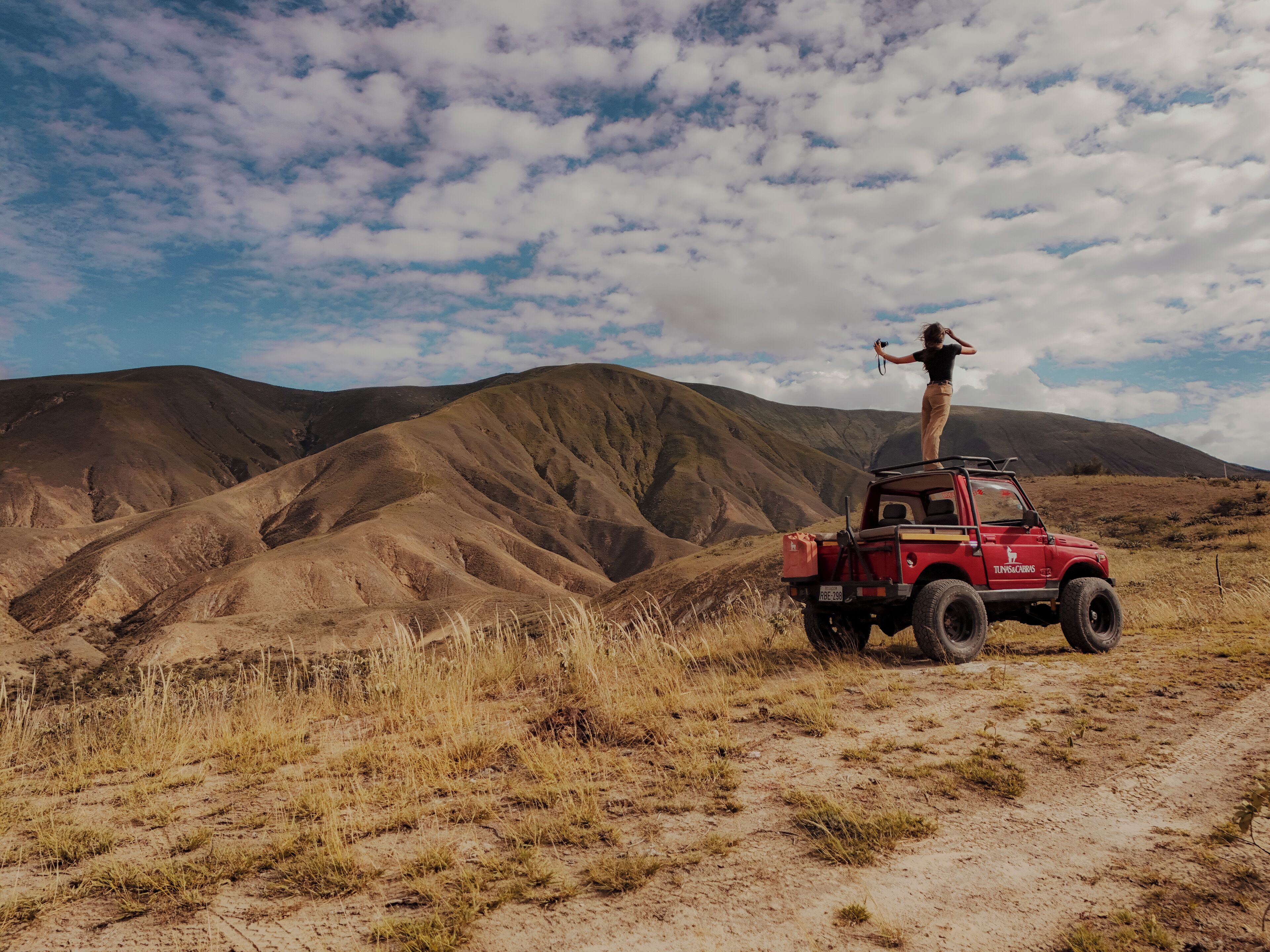 A red off-road vehicle parked on a dirt path with a person standing on top, surrounded by a mountainous landscape.