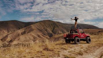 A red off-road vehicle parked on a dirt path with a person standing on top, surrounded by a mountainous landscape.