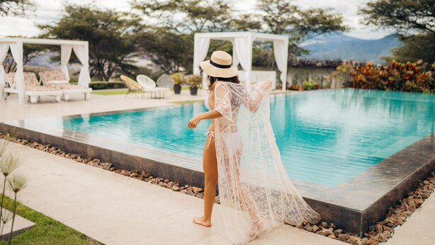 Ein Poolbereich mit einer Person in weißem Kleid und Hut, einer weißen Laube und Blick auf Bäume und Berge im Hintergrund.