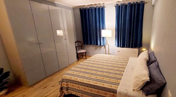 A bedroom with a bed, a chair, a wardrobe, and a nightstand with a lamp. - Freshly Renovated Apartment in the Historic Centre close to the Duomo (Siena)