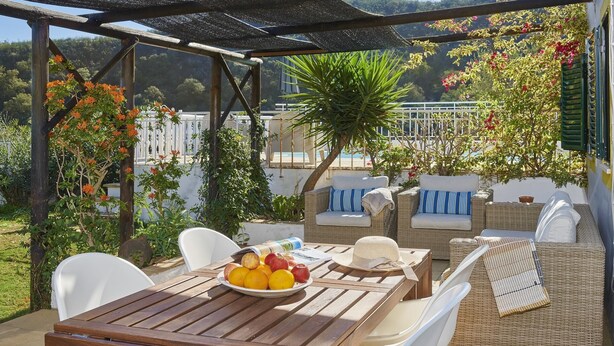 An outdoor patio with wicker furniture, a wooden table with fruits, and a view of a garden and hills. - Charming Rural Retreat, A/C, Private Pool & Beautiful Views - 11km from the Coastal Town of Tavira! (Tavira)
