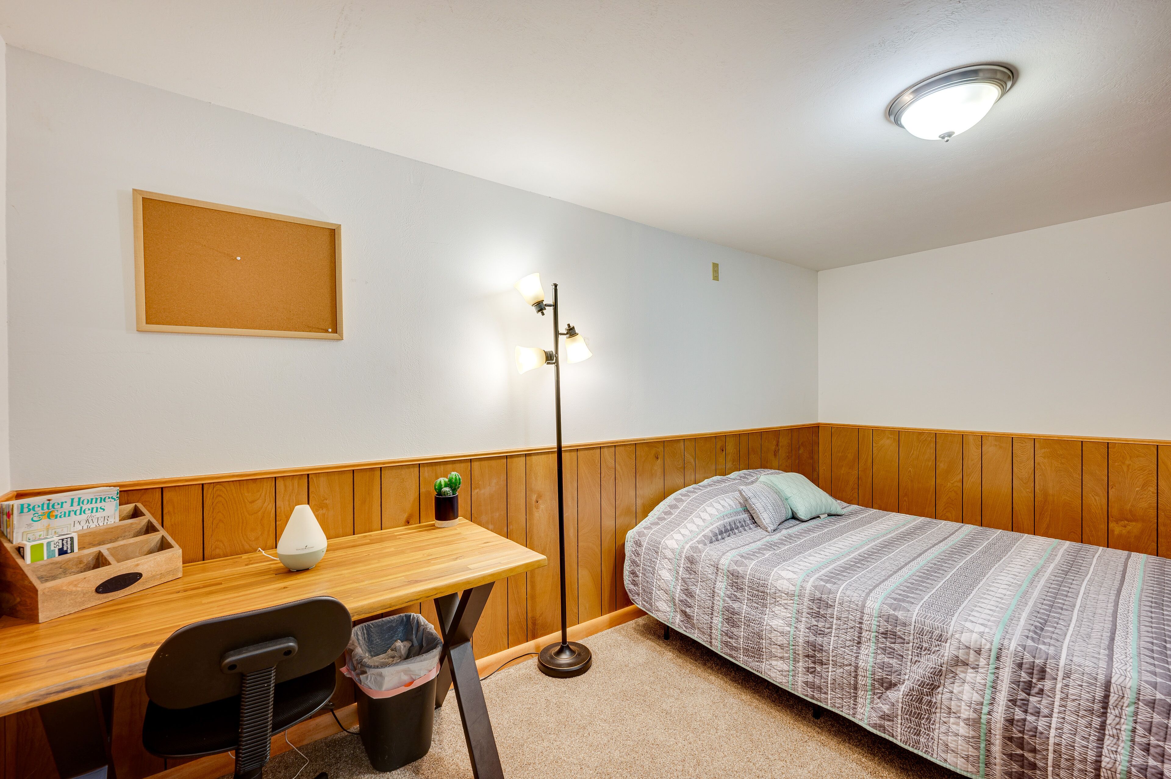 Ein kleiner, ordentlicher Raum mit einem Holztisch, einem Bett mit gemustertem Überwurf, einer Stehlampe und einem gerahmten Bild an der Wand.