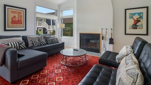 Un salón moderno con un sofá gris, un sofá negro, una mesa de centro y una alfombra roja estampada. Hay dos ventanas, una chimenea y dos cuadros en las paredes.