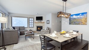 A modern living room with a fireplace, a dining area with a table set for four, and a large window offering a view of the outdoors.