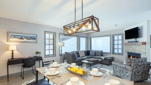 A modern living room with a dining area, large windows, a fireplace, and a mounted TV.