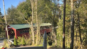 A covered outdoor seating area with a wooden bench and a table, surrounded by trees and a red building in the background. - Fly fishing, tubing, biking and shopping are steps away! (Sunderland)