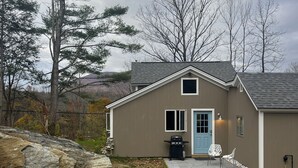 A single-story house with a brown exterior, a blue door, and a small porch with two white chairs. - Fly fishing, tubing, biking and shopping are steps away! (Sunderland)