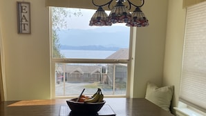 A dining area with a dark wooden table, a bowl of fruit, and a view of a lake and houses through a large window. - The Spot on Sproat Lake (Port Alberni)