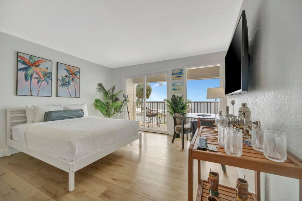 A bedroom with a bed, a TV, a balcony with plants, and a table with glasses.