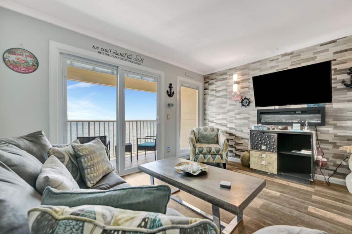 A living room with a sofa, armchair, and coffee table. There is a TV mounted on the wall, a fireplace, and a balcony with a view.