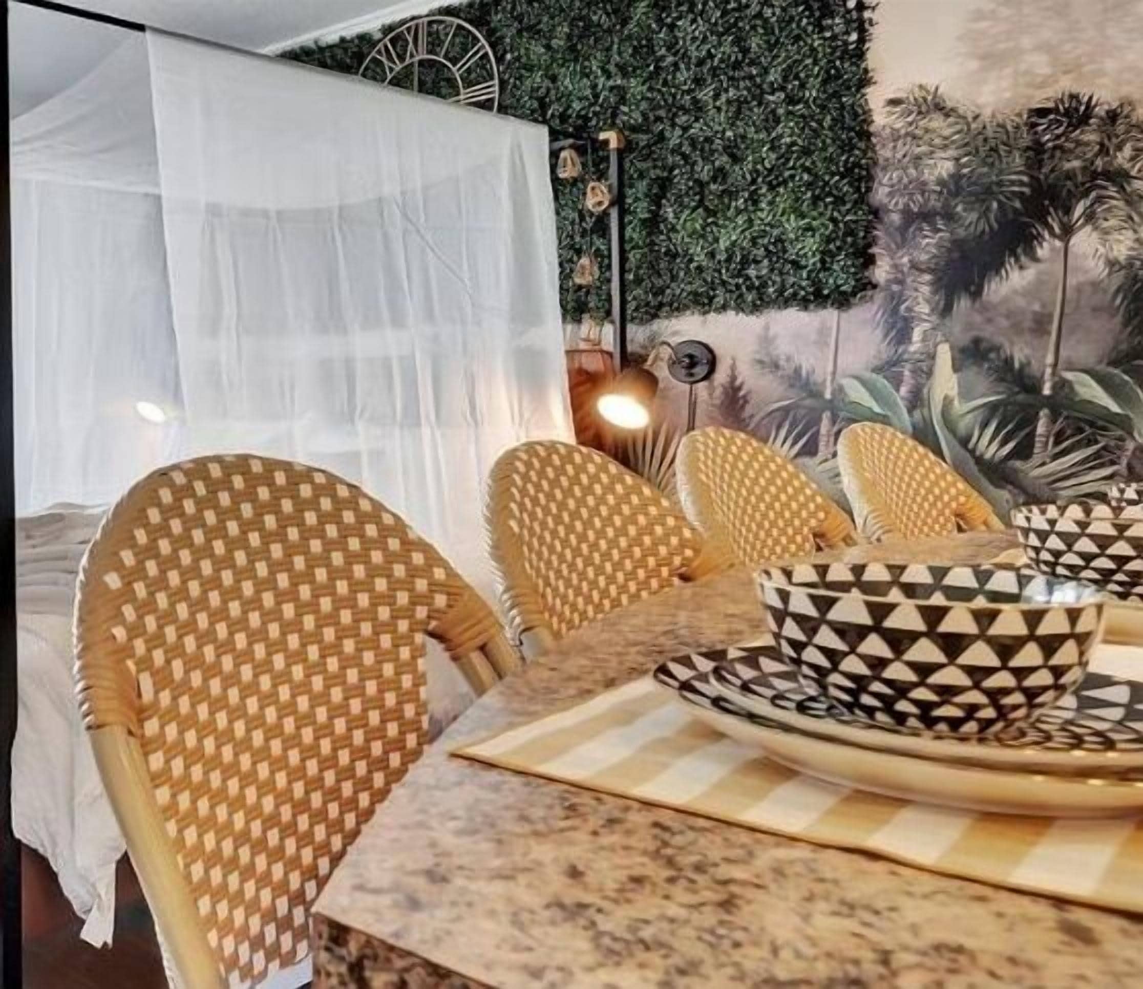 A dining area with wicker chairs, a table with a patterned table runner, and a wall with a green leafy mural.