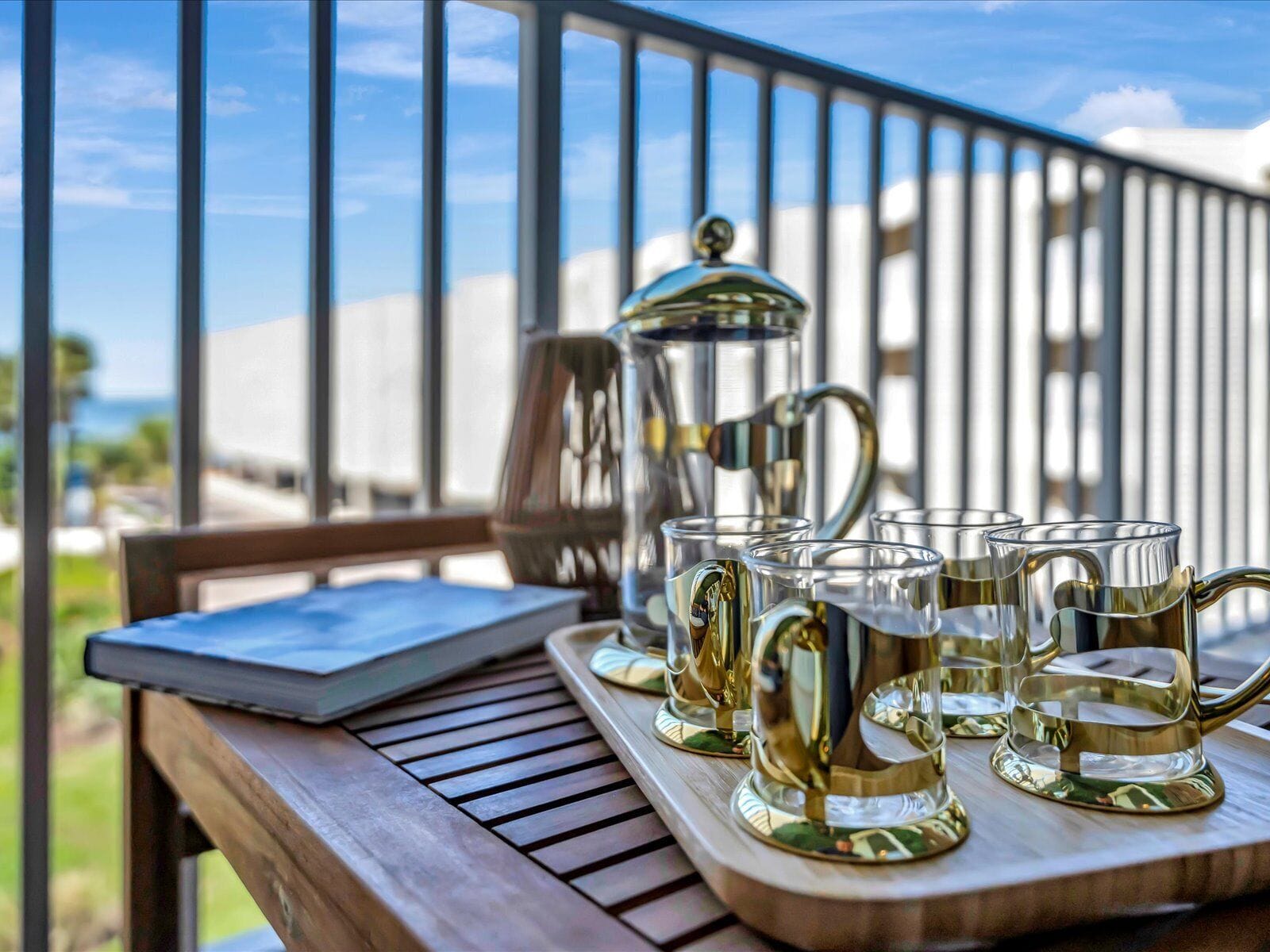 Ein Holztisch mit einer Tablett, auf dem eine Kaffeekanne und mehrere Gläser stehen, auf einem Balkon mit Geländer und Blick aufs Meer.