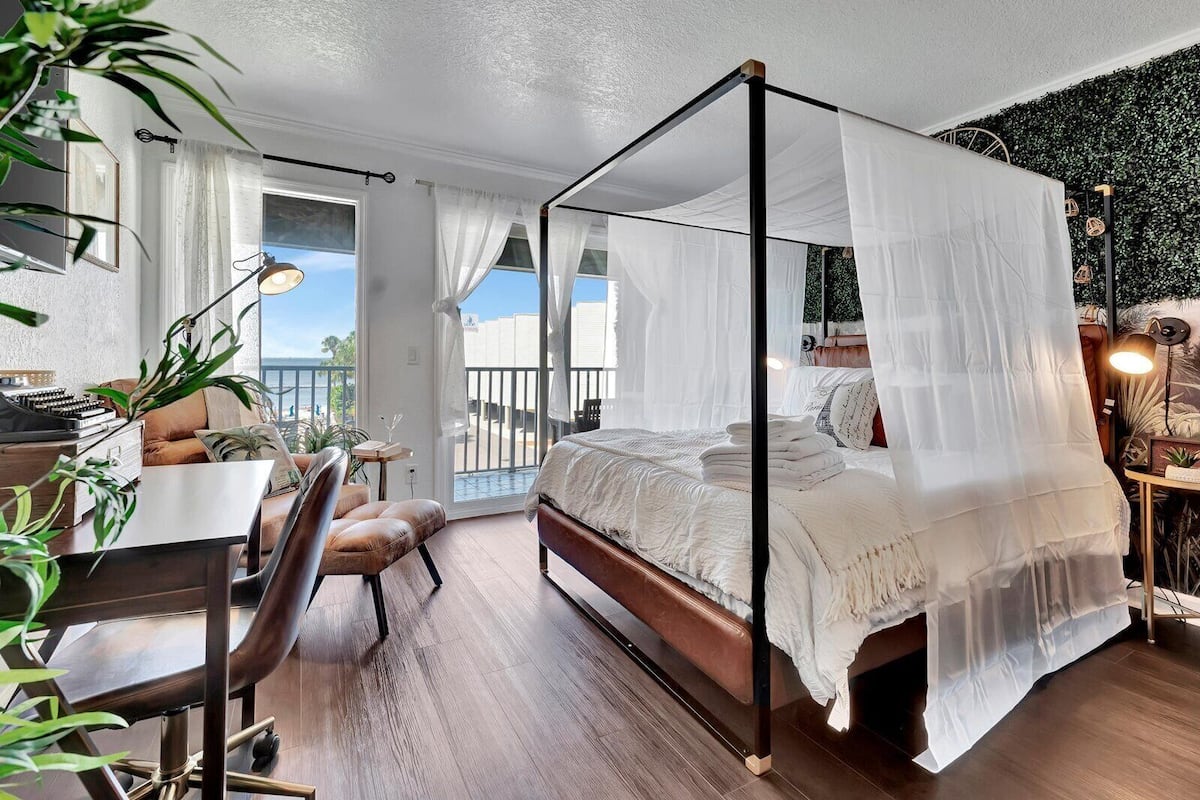 A four-poster canopy bed with a view of the outdoors, a desk with a chair, and a balcony with a railing.