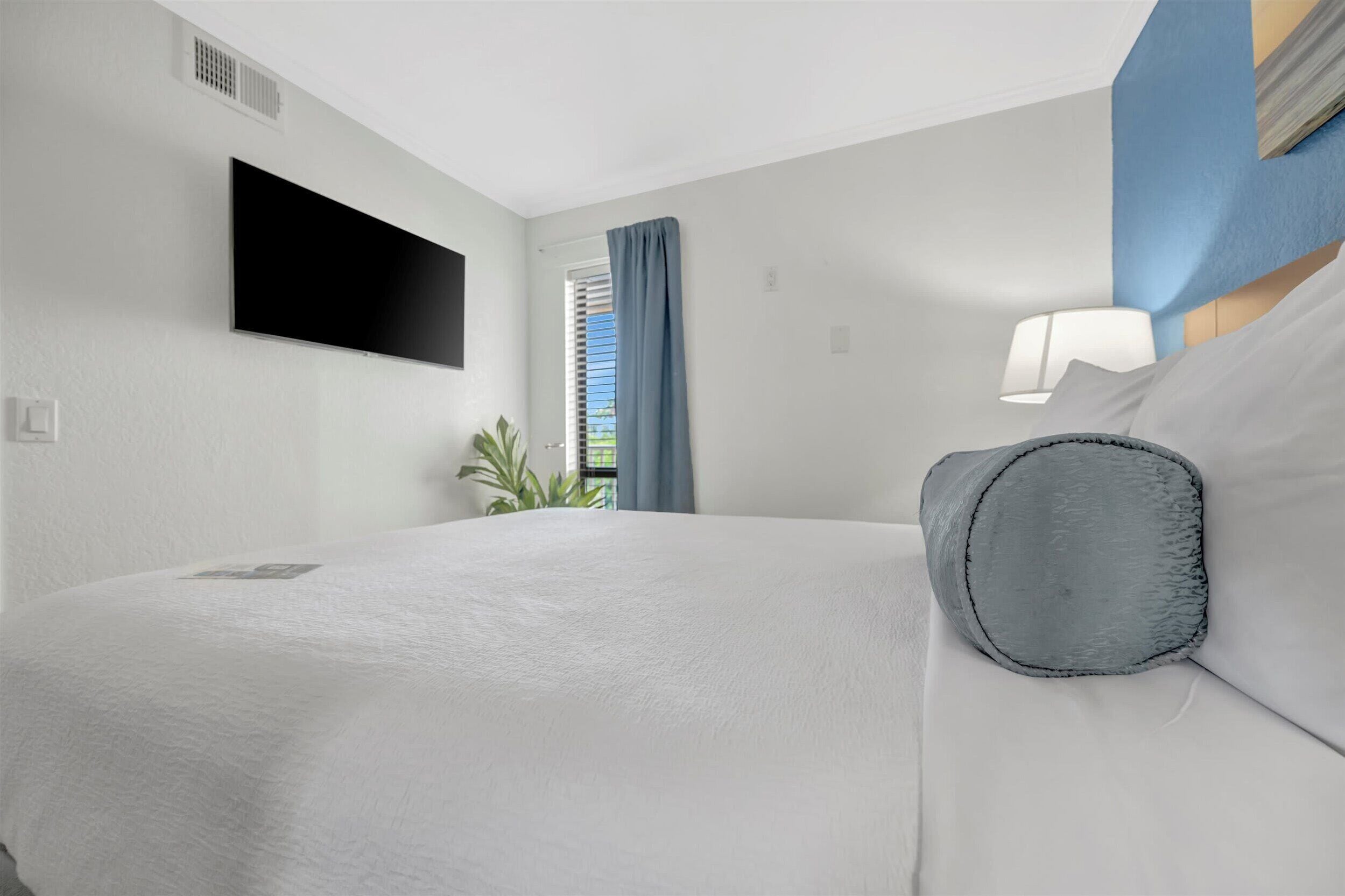 A hotel room with a bed, a flat-screen TV mounted on the wall, a window with blue curtains, and a bedside lamp.
