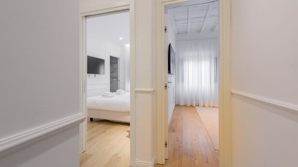 Un pasillo que da a un dormitorio con cama, espejo y ventana con cortinas.