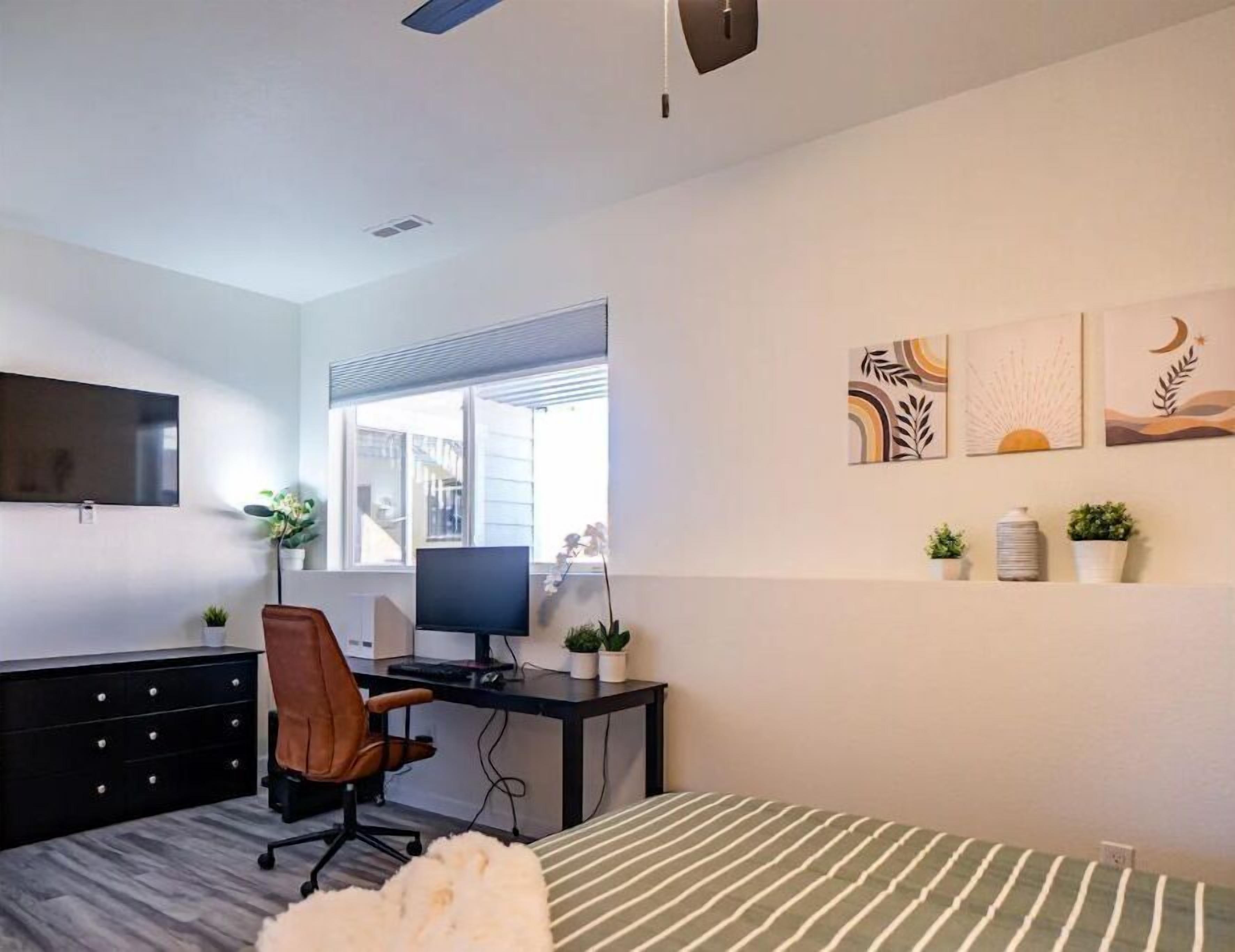 Un dormitorio con cama, escritorio con computadora, silla, televisor y obras de arte en la pared.
