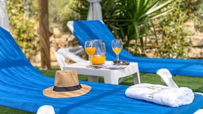 Een ligbed met een blauwe hoes, een strohoed, een handdoek en twee glaasjes sinaasappelsap op een tafel.