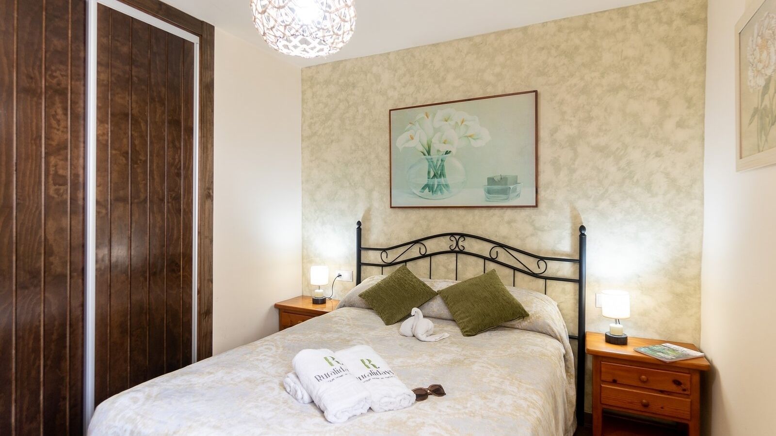 Ein ordentlich bezogenes Bett mit weißen Leinen und grünen Kissen, ein gerahmtes Blumenbild an der Wand und eine Holztür links.