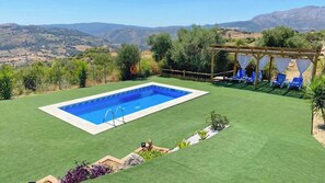 A rectangular swimming pool with a wooden deck and lounge chairs, surrounded by green artificial grass and a view of distant hills. - Casa Salysol Algatocín by Ruralidays (Málaga)