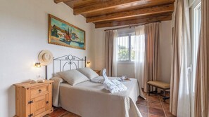 A bedroom with a bed, bedside table, lamp, painting, window, curtains, and a bench. - Casa La Bimbre Algatocín by Ruralidays (Málaga)