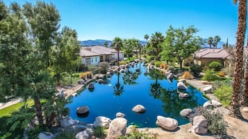 Un área de piscina tranquila con una piscina azul cristalina, rodeada de rocas y diferentes árboles, y casas al fondo.