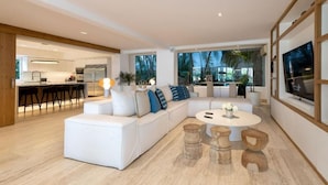 Ein modernes Wohnzimmer mit einem weißen Sofa, einem runden Couchtisch und Holzschemeln. Im Hintergrund ist eine Küche mit weißen Schränken und einer Bar zu sehen.