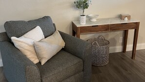 A grey sofa with cushions, a small white table with a vase of green plants, a round mirror, and a wicker basket.