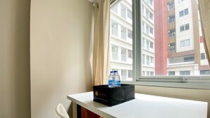 A hotel room with a white desk, a black box labeled 'Travelio', and a white chair. A window reveals a building with balconies and a red pillar. - Great Choice Studio Apartment LRT City Jatibening (Bekasi)
