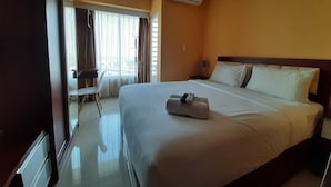 A hotel room with a large bed, an air conditioning unit, a chair, and a view of the city through the window. - Comfort Living Studio Apartment At Grand Kamala Lagoon Near Shopping Center (Bekasi)