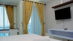 A hotel room with a bed, a TV mounted on the wall, a window with curtains, and a small shelf with a remote control. - Modern And Comfy Studio At Grand Dhika City Apartment (Bekasi)