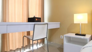 A hotel room with a desk, chair, bedside table, lamp, and curtains. - Comfortable Studio Apartment Grand Dhika City (Bekasi)
