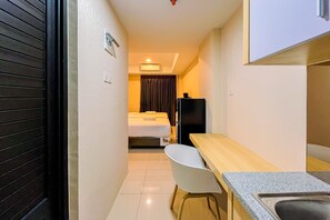 Een compacte hotelkamer met een bed, bureau, stoel, koelkast en een klein wastafeltje.