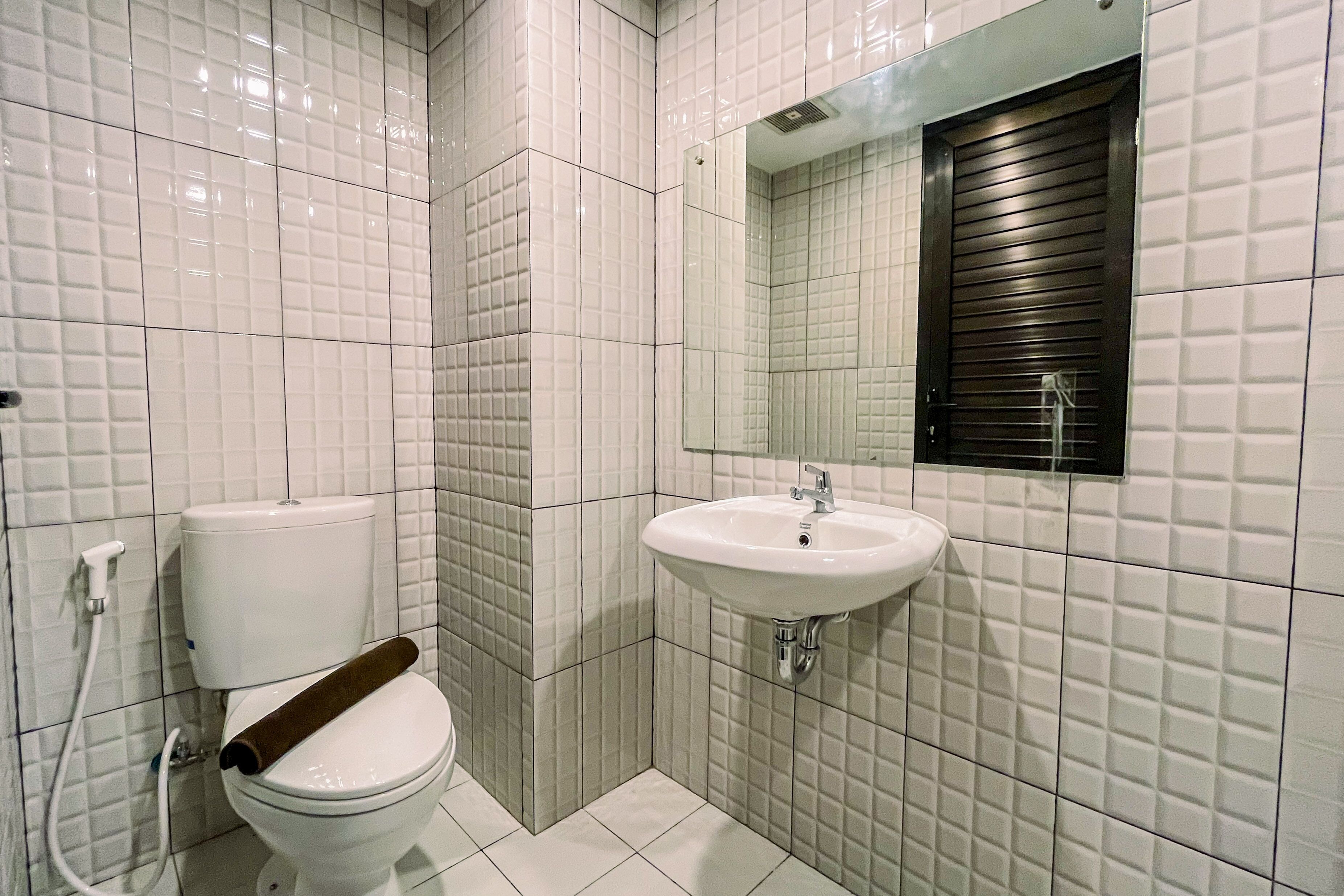 Un baño con paredes de azulejos blancos, un inodoro con asiento de madera, un lavamanos blanco y un espejo con un ventanal arriba.