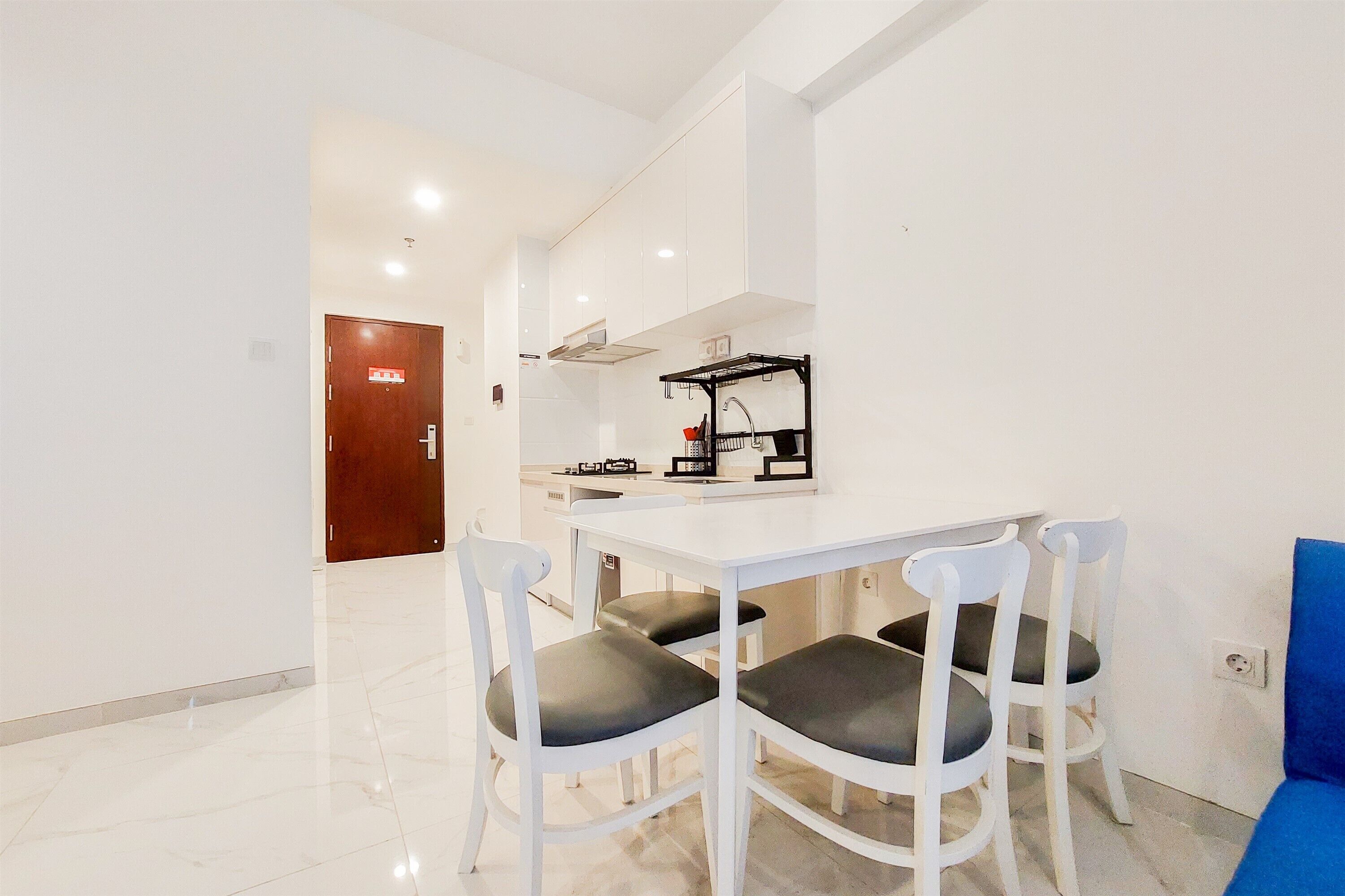 A modern kitchen with white cabinets, a black hanging rack, and a white countertop. There are three white chairs with black cushions around a white table.