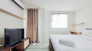 A hotel room with a bed, a TV on a wooden stand, a window with curtains, and an air conditioning unit. - Homey Studio At Bintaro Icon Apartment (South Tangerang)
