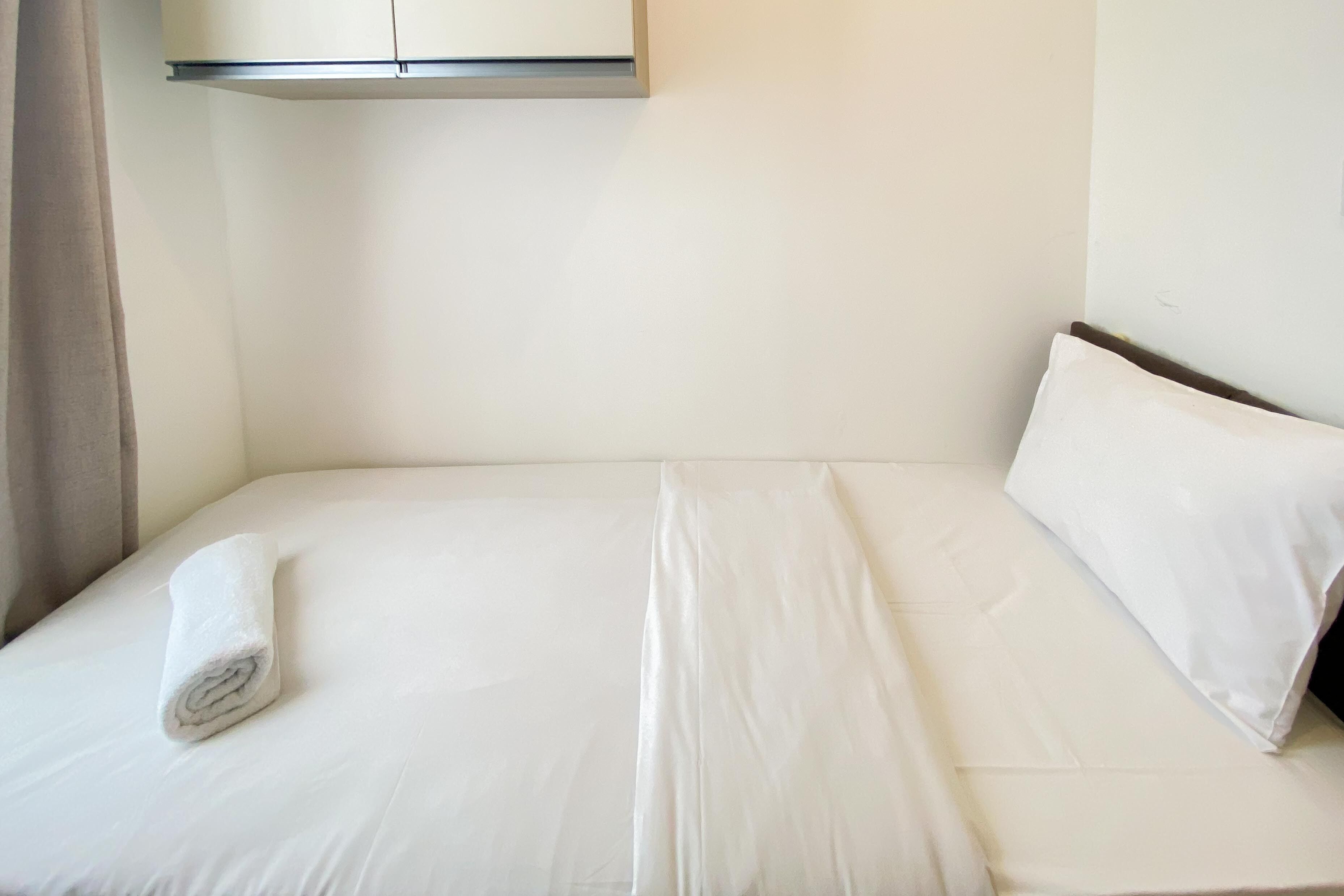 A neatly made bed with white linens and a rolled towel, a white wall, and a ceiling-mounted light fixture.