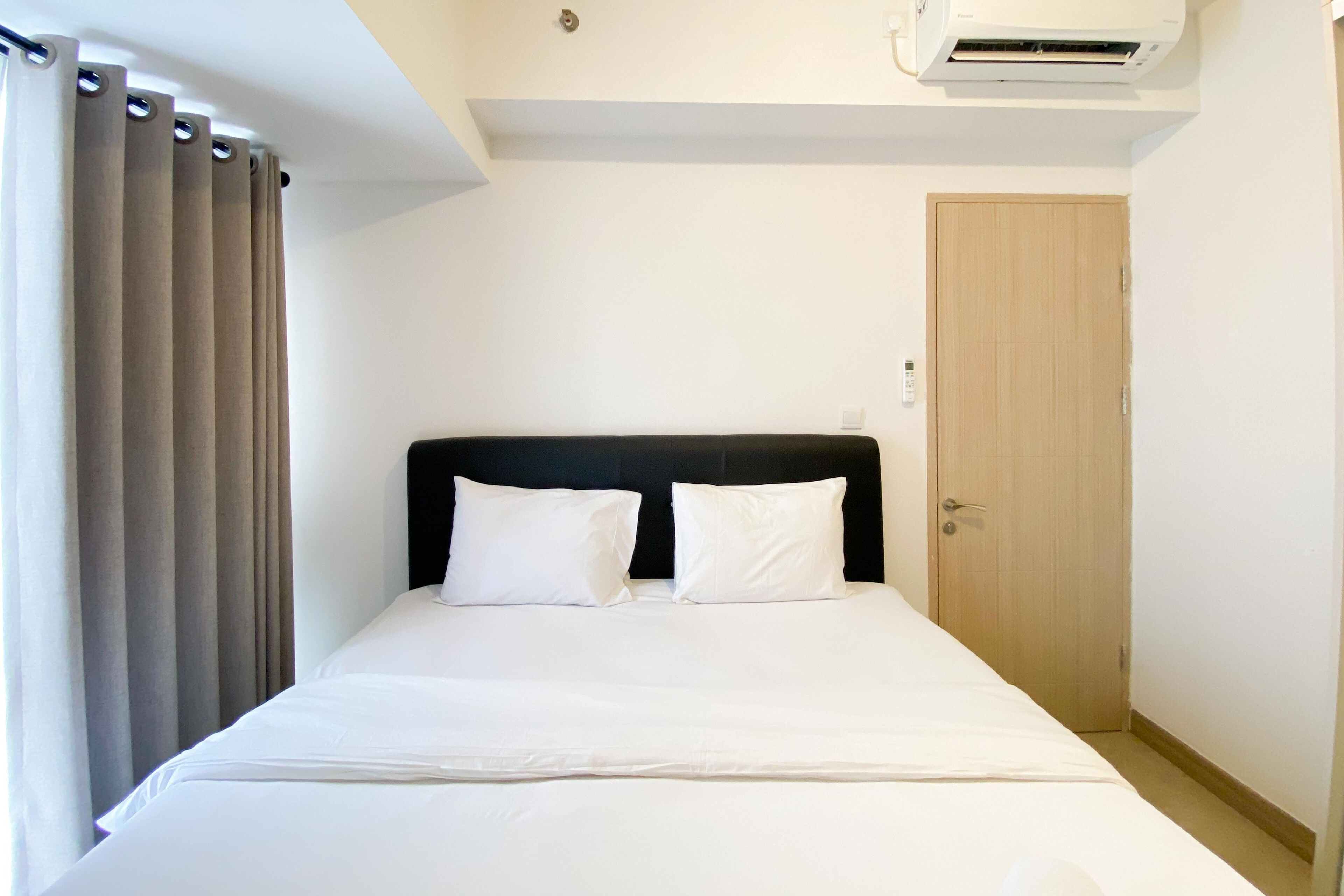 Cama bem arrumada com lençóis brancos e duas almofadas, uma porta de madeira à direita e um aparelho de ar condicionado sobre a cama.
