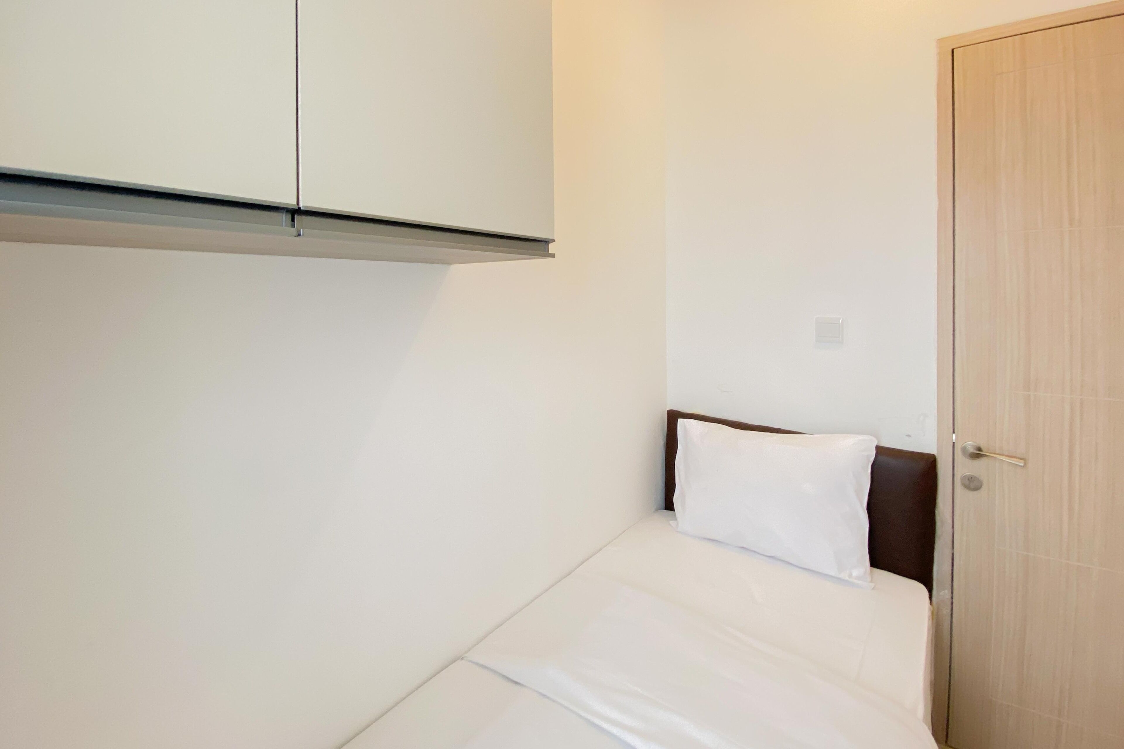 Une petite chambre avec un lit simple, une tête de lit en bois, un oreiller blanc et une porte fermée.