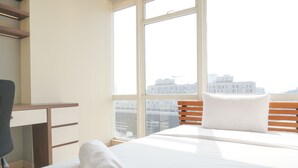 A hotel room with a bed, a desk, and a view of the cityscape through large windows.