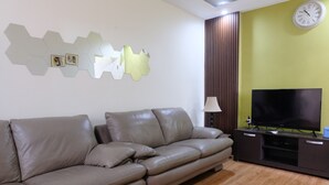 A modern living room with a brown leather sofa, a flat-screen TV on a dark wooden entertainment unit, and a wall clock. - Spacious And Modern 3Br Apartment At Gateway Ahmad Yani Cicadas (Bandung)