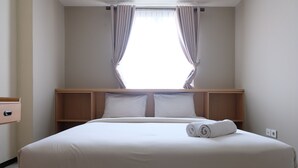 Un lit bien fait, avec des draps blancs et une serviette enroulée, une tête de lit en bois et une fenêtre avec des rideaux.