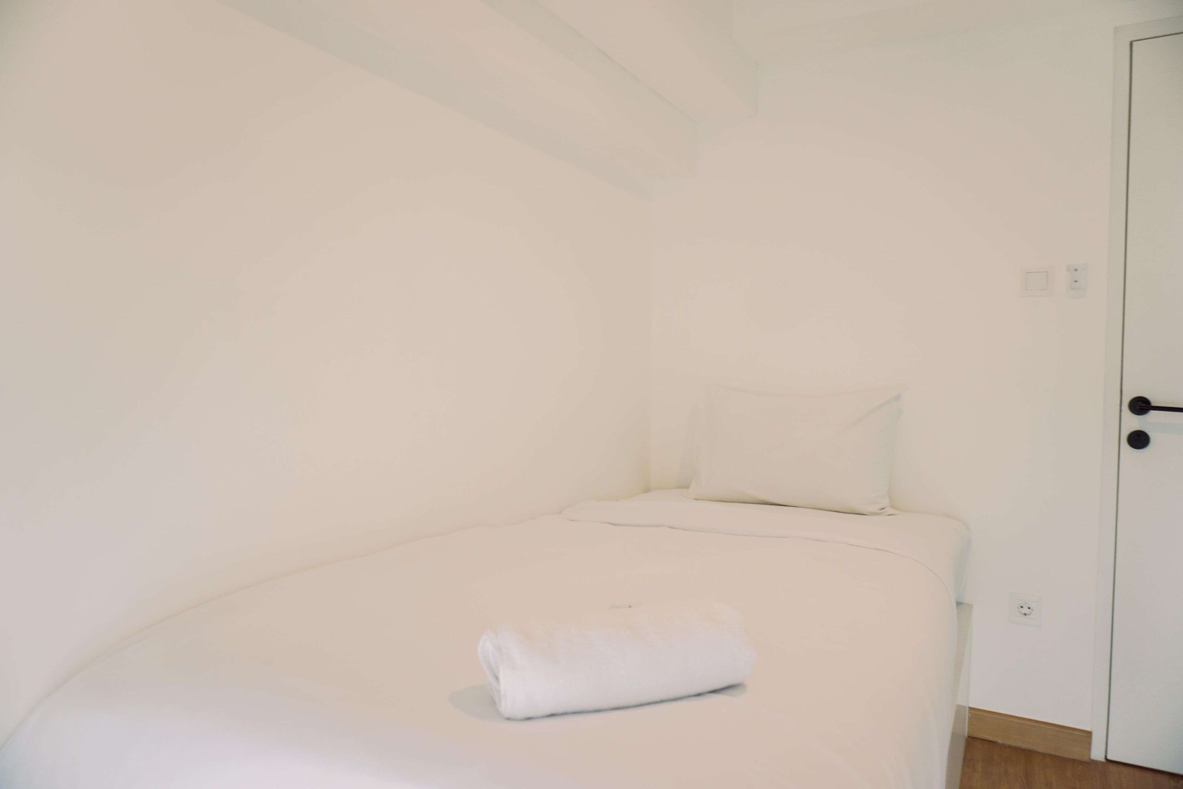 A small, white room with a single bed, a white pillow, and a folded white towel.
